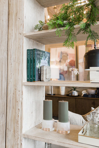 Two green ribbed pillar candles on whitewashed wooden bookshelf featuring other decor pieces. 
